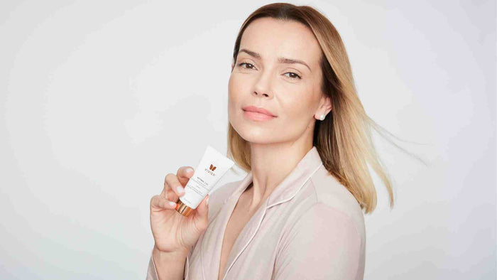 Person holding a Vivier Retinol 1.0% cream tube while looking toward the camera against a soft neutral background.