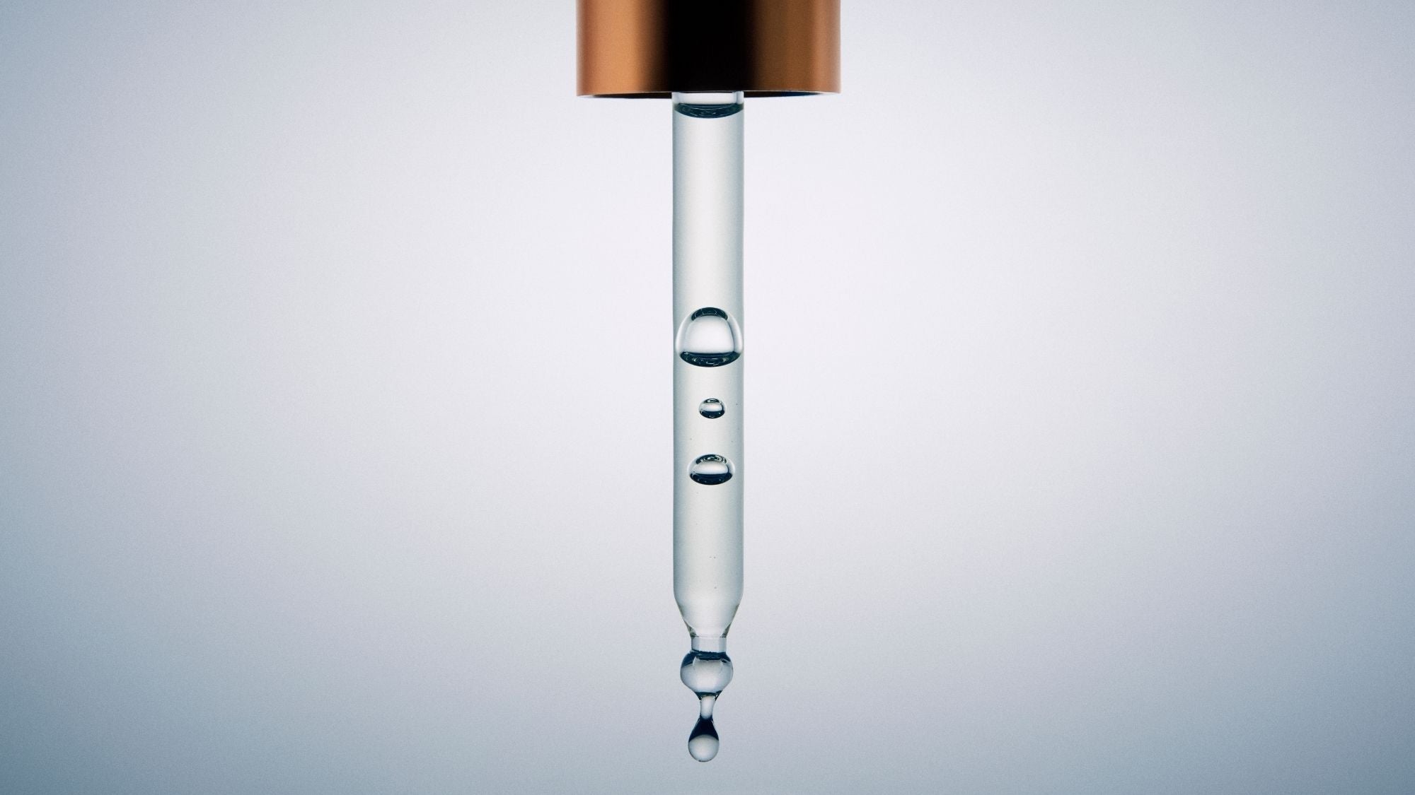 Close-up of a glass serum dropper containing clear skincare serum with suspended droplets against a soft grey background.